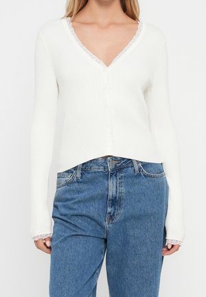 Femme portant un cardigan blanc côtelé boutonné avec une bordure en dentelle et un jean bleu taille haute, debout les mains détendues sur les côtés.