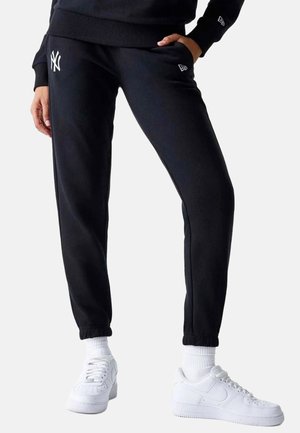 Personne portant un pantalon de survêtement noir avec le logo blanc des New York Yankees, des chaussettes blanches et des baskets blanches, debout avec une main dans la poche.