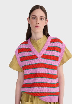 Jeune femme portant un gilet tricoté rayé rose et rouge sur une robe moutarde à manches courtes boutonnée, posant devant un fond clair uni.