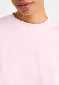 Felpa rosa chiaro realizzata in morbido tessuto, con scollatura a girocollo e logo Carhartt in rilievo sul petto. Tessuto liscio e vestibilità comoda.