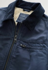 Veste bleu marine avec col en velours côtelé, fermeture éclair frontale, poche poitrine zippée et doublure polaire beige visible au niveau du col.