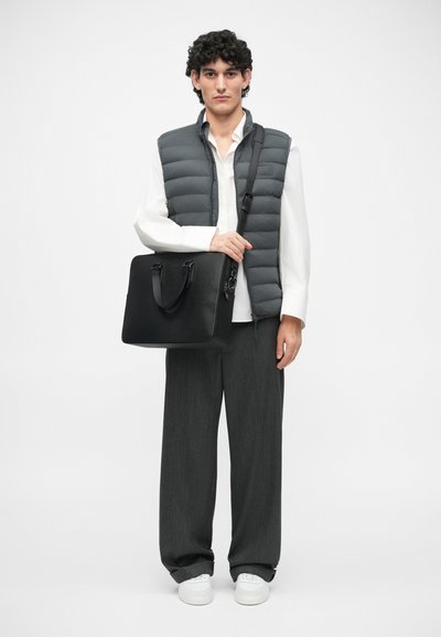 Sac à mallette en cuir noir avec poignées supérieures et bandoulière. Le modèle porte un gilet rembourré gris, une chemise blanche et un pantalon large gris. Baskets blanches.