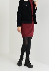 Chaqueta de pana negra con cuello de borrego, vestido burdeos, medias negras y botas negras con cordones y suelas gruesas.