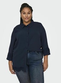 Camicia blu navy con bottoni e colletto, caratterizzata da dettagli di pizzo intricati sui polsini e sull'orlo. Indossata con jeans in denim scuri.