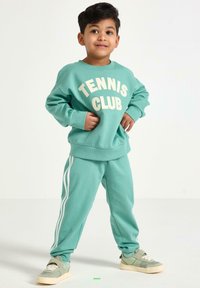 Miętowa bluza z kapturem z nadrukiem "Tennis Club" oraz pasujące spodnie dresowe z białymi paskami po bokach, w zestawie z jasnemi zielonymi sneakersami.
