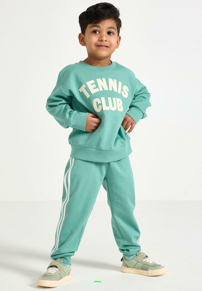Miętowa bluza z kapturem z nadrukiem "Tennis Club" oraz pasujące spodnie dresowe z białymi paskami po bokach, w zestawie z jasnemi zielonymi sneakersami.