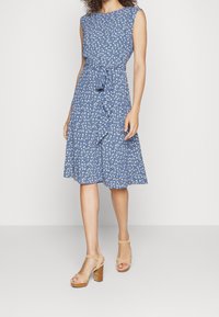 Blå blommönstrad klänning med figursydd midja, ärmlös design och knälång kjol. Tillsammans med beige klumpklackade sandaler.