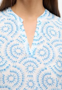 Blusa leggera bianca con motivo tie-dye blu. Caratterizzata da scollatura a V e una texture morbida e liscia.