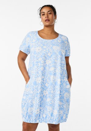 Kvinde iført en lyseblå knælange kjole med hvide blomsterprint, korte ærmer og hænderne i lommerne, foran en hvid baggrund.