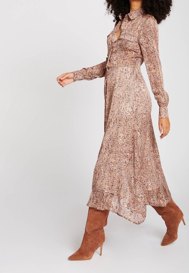 Femme marchant dans une robe midi à manches longues, boutonnée et à motifs, associée à des bottines marron en daim à talons, sur un fond uni.