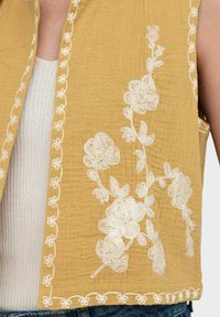 Senapsgul väst med vit blommig broderi längs kanterna och på bröstet. Lätt textur, öppen framdesign, utan ärmar, lagd över en ribbad topp.