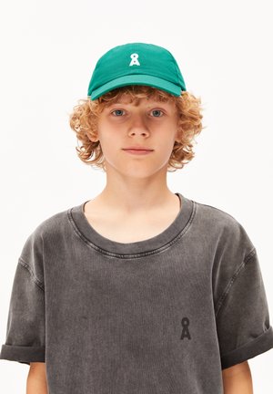 Groene pet met een logo aan de voorkant en een gebogen rand, gecombineerd met een loszittend, grijs t-shirt met een klein logo op de borst.