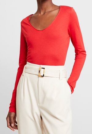 Personne portant un haut rouge moulant à manches longues avec un décolleté rond, rentré dans un pantalon blanc taille haute avec une large ceinture et une boucle rectangulaire.