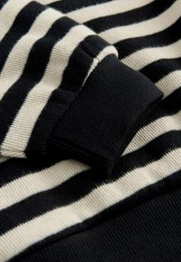 Tissu tricoté noir et crème à rayures avec une surface texturée. Poignet noir ajusté montrant une couleur contrastante et des détails côtes. Rayures horizontales visibles.