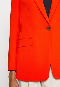 Červený pánský oblekový blazer se zapínáním na jeden knoflík, klopovými límci, boční kapsou a texturovanou tkaninou. Stylizováno s kalhotami světlejší barvy.