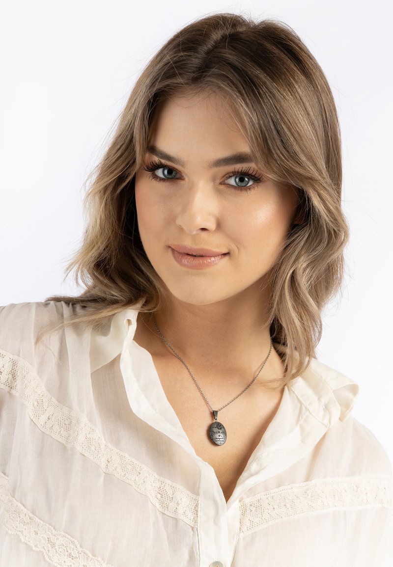 Collana con pendente d'argento con dettagli incisi, su una catena sottile, indossata sopra una blusa chiara e texturizzata con accenti in pizzo.