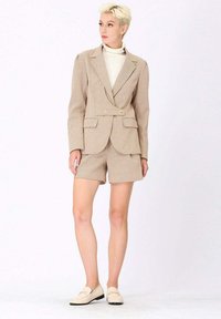 Blazer à carreaux beige avec deux boutons, associé à un short assorti. Le modèle porte un col roulé blanc et des mocassins de couleur claire.