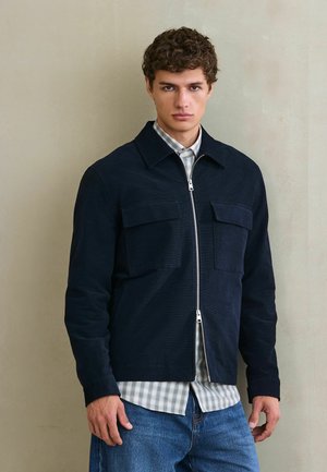 Junger Mann mit lockigem Haar, der eine dunkelblaue Jacke mit Reißverschluss über einem hell karierten Hemd und blauen Jeans trägt, steht vor einer schlichten beigen Wand.