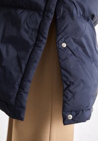 Jūras zilā pūkainā jaka ar spīdīgu virsmu, pogu aizdari un vieglu, quilted struktūru, daļēji segot brūnu apģērbu.