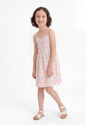 Jeune fille souriante portant une robe pastel sans manches avec des motifs de cœurs multicolores et des sandales blanches, debout sur un fond blanc.