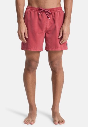 Mann trägt rote Badehose bis zur Mitte des Oberschenkels mit Kordelzug in der Taille und steht barfuß vor einem schlichten weißen Hintergrund.