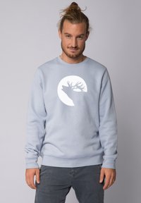 Lichtblauwe sweatshirt van zacht materiaal, met een witte silhouet van een hert tegen een cirkelvormig ontwerp op de borst. Ribbelacinten en zoom.