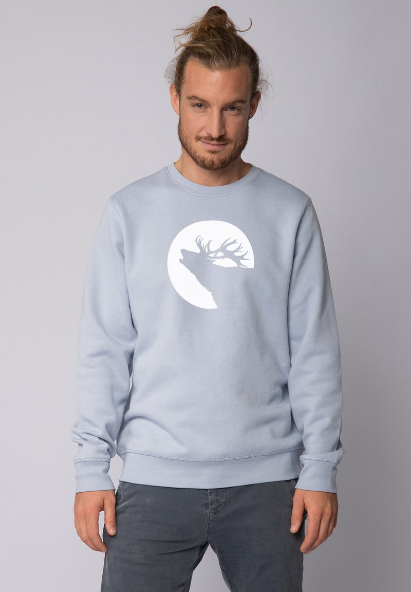 Lichtblauwe sweatshirt van zacht materiaal, met een witte silhouet van een hert tegen een cirkelvormig ontwerp op de borst. Ribbelacinten en zoom.
