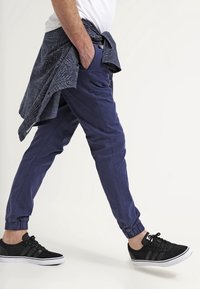 Man loopt, gekleed in marineblauwe joggingsbroek, zwarte sneakers, een wit t-shirt en een met patroon versierde jas om zijn taille gebonden, tegen een effen lichte achtergrond.