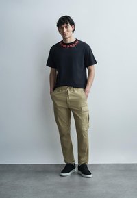 Jeune homme debout les mains dans les poches, portant un t-shirt noir à motif graphique, un pantalon cargo beige et des baskets noires, sur un fond gris uni.