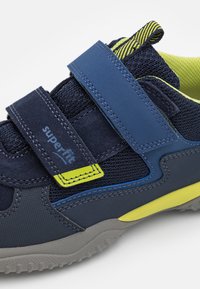 Granatowe i limonkowe buty sportowe z siateczkową i zamszową cholewką, rzepami oraz teksturowaną szaro-rubberową podeszwą.