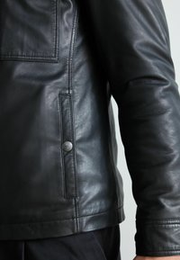 Gros plan d’une personne portant une veste en cuir noire avec des coutures visibles et une poche latérale à bouton-pression.