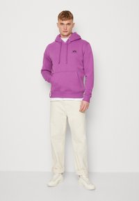 Felpa con cappuccio viola, dotata di tasca frontale e cordino. Abbinata a pantaloni beige arrotolati e sneakers bianche. Visibile un branding minimal.