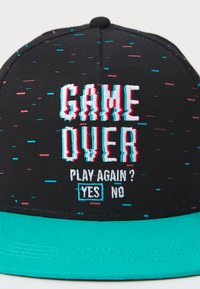 Svart caps med en turkise skjerm; har broderte tekster: "GAME OVER" og "PLAY AGAIN? YES NO" i blått og rosa. Teksten har en glitch-effekt.