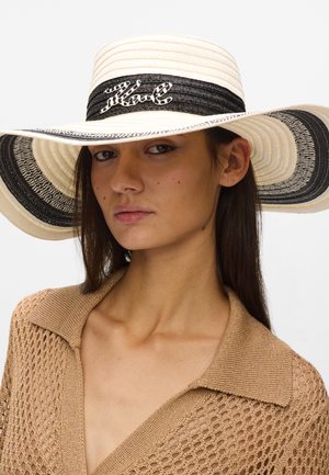 Ženska, ki nosi širokokrajni slamnik s črnimi in belimi trakovi ter bež pleteno majico z V-izrezom in ovratnikom.