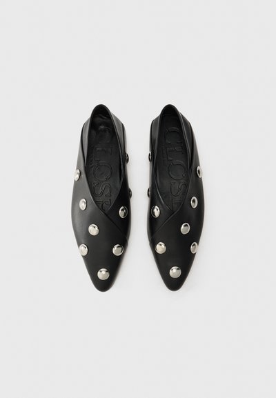 Chaussures en cuir noir avec un bout pointu, présentant un motif en croix et des ornements circulaires en argent. Texture lisse et style à enfiler.