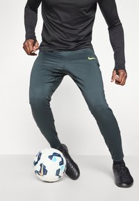 Tamnozelene atletske hlače uskog kroja s žutim Nike logom, u kombinaciji s crnom majicom dugih rukava i crnim sportskim cipelama pored nogometne lopte.
