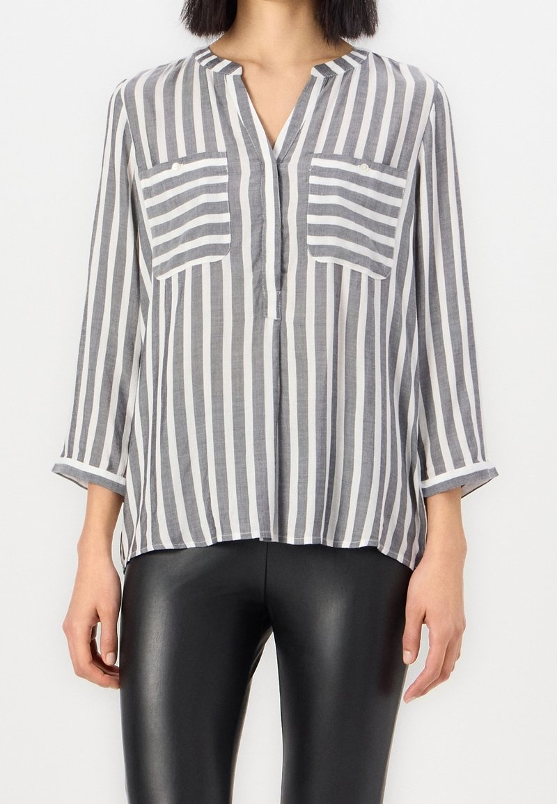 Blouse légère à rayures grises et blanches, avec un col en V, deux poches poitrine et des manches trois-quarts. Associée à un legging noir.