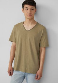 Olijfgroene t-shirt met V-hals van lichtgewicht, gestreept materiaal met korte mouwen. Gecombineerd met lichtblauwe spijkerbroek, weergegeven tegen een neutrale achtergrond.