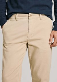 Pantaloni in cotone beige con una superficie liscia, dotati di chiusura con bottone e vestibilità classica. Gamba leggermente affusolata con tasche laterali.