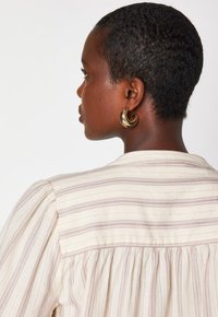 Profilo chiuso di una persona con capelli corti e ricci, che indossa una maglietta a righe crema e grandi orecchini a cerchio dorati su uno sfondo bianco.