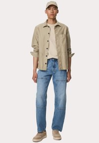 Beige katoenen shirtjack met zwarte knopen over een beige t-shirt, gecombineerd met lichtblauwe denim jeans en beige sneakers.