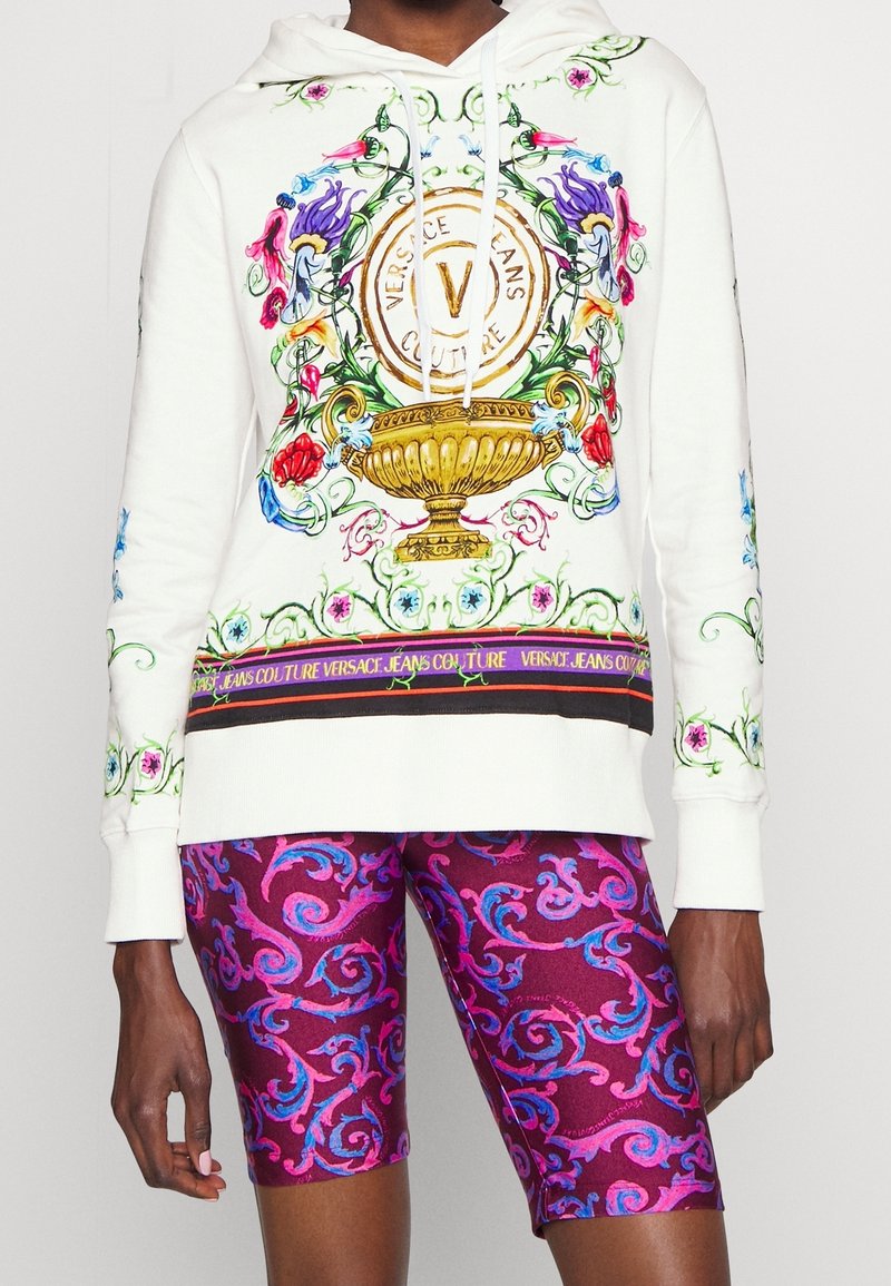 Persona con sudadera blanca con un diseño ornamentado de flores y una urna dorada, y pantalones cortos con un patrón en tonos púrpura y rosa.