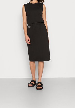 Mujer con vestido midi negro sin mangas con pequeño texto impreso en el pecho y área del bolsillo, combinado con sandalias negras de punta abierta.