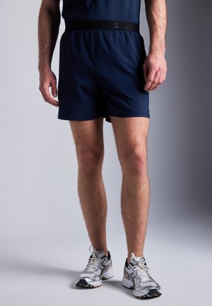 Marina träningsshorts med elastisk midja, slät textur och sidoflikar. Kombineras med ljusgrå och svarta löparskor.