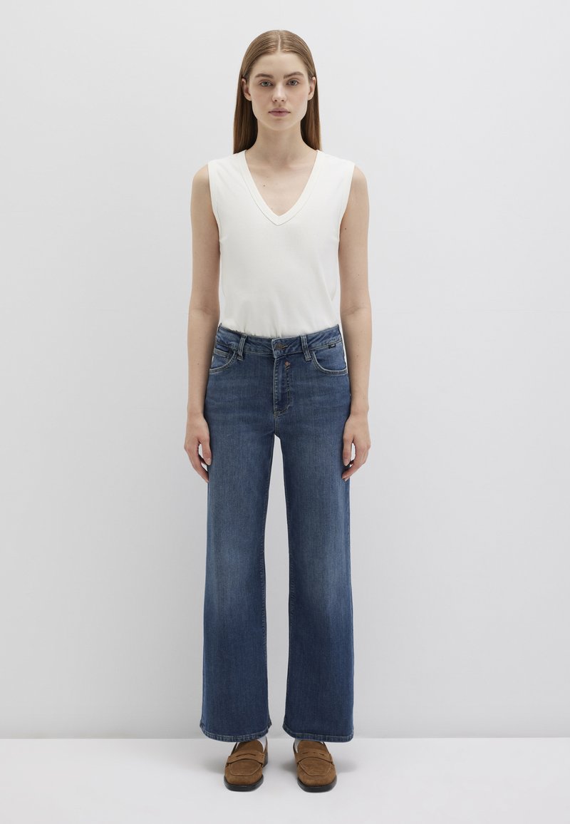 White sleeveless top with a v-neck, paired with high-waisted wide-leg blue jeans. Model wearing brown slip-on shoes. Plain background.