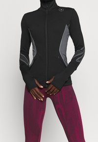 Black fitted jacket with gray side panels, high collar, full zip, and thumbholes. Accompanied by patterned maroon leggings.
