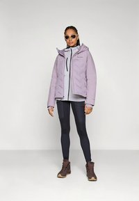 Lyse lilla dunjakke med hætte og lynlås foran, båret over en lysegrå top. Kombineret med sorte leggings og brune vandrestøvler.