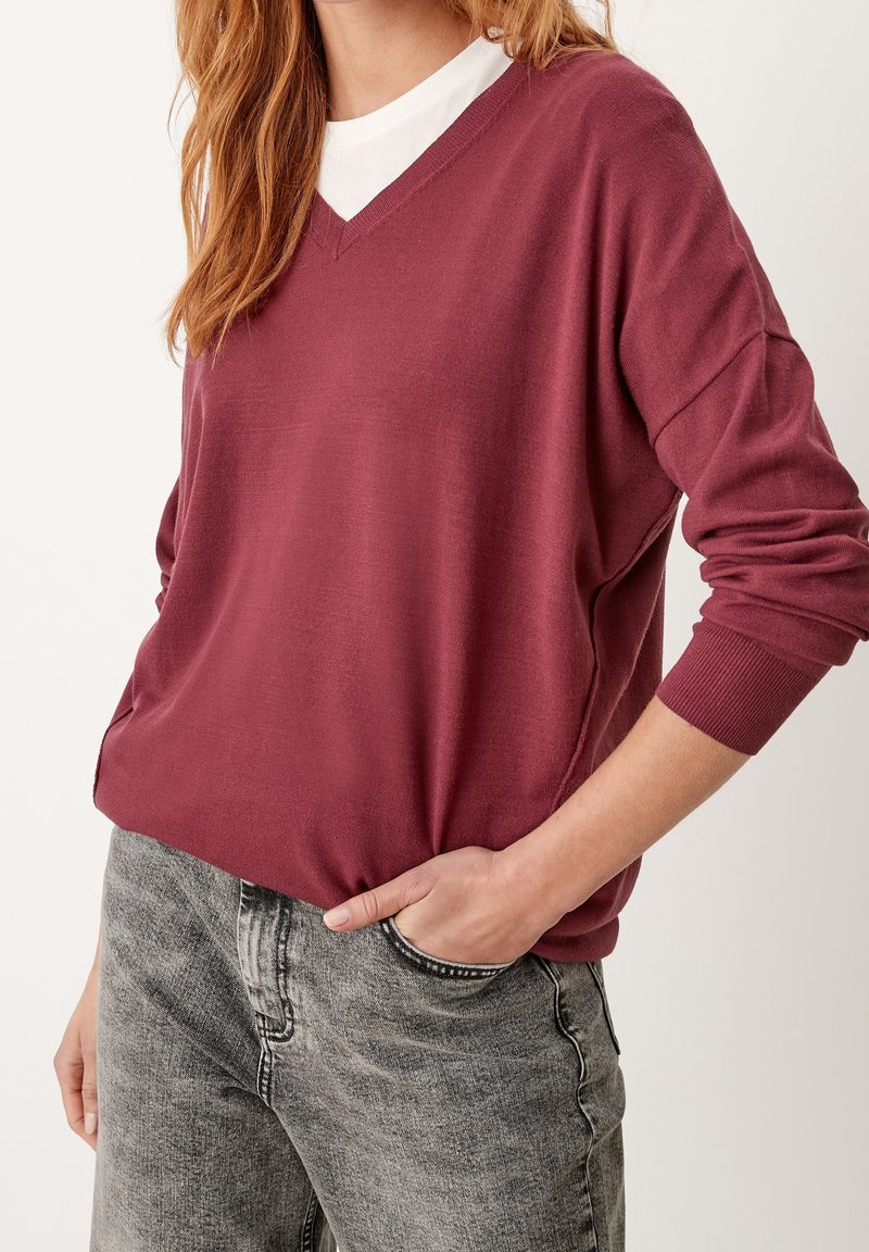 Femme portant un pull col en V bordeaux par-dessus un top blanc, une main dans la poche d'un jean gris délavé, sur un fond uni.