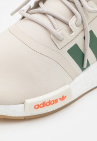 Beige sportskor med övre del i slätt tyg, gröna detaljer och vit mellansula. Har orange logotypdetalj och texturerad yttersula.