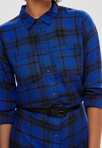 Niebieska flanelowa koszula w kratę z czarnymi akcentami, posiadająca dwie kieszenie na piersi, kołnierzyk zapinany na guziki i noszona z czarnym paskiem.